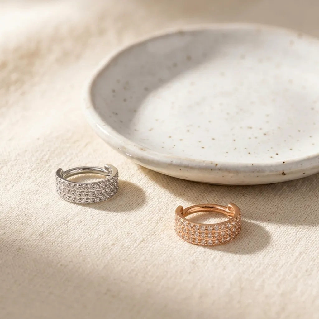 Two slim pave band rings, one silver-toned and one rose gold-toned, displayed on a textured beige fabric next to a speckled ceramic dish.