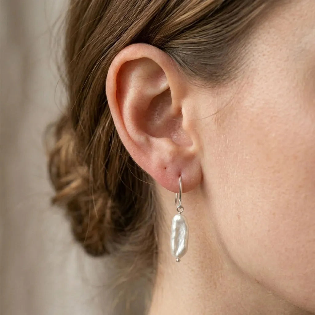 A detailed close-up shot of a woman with her hair in a bun, wearing a single baroque pearl drop earring with a silver hook.