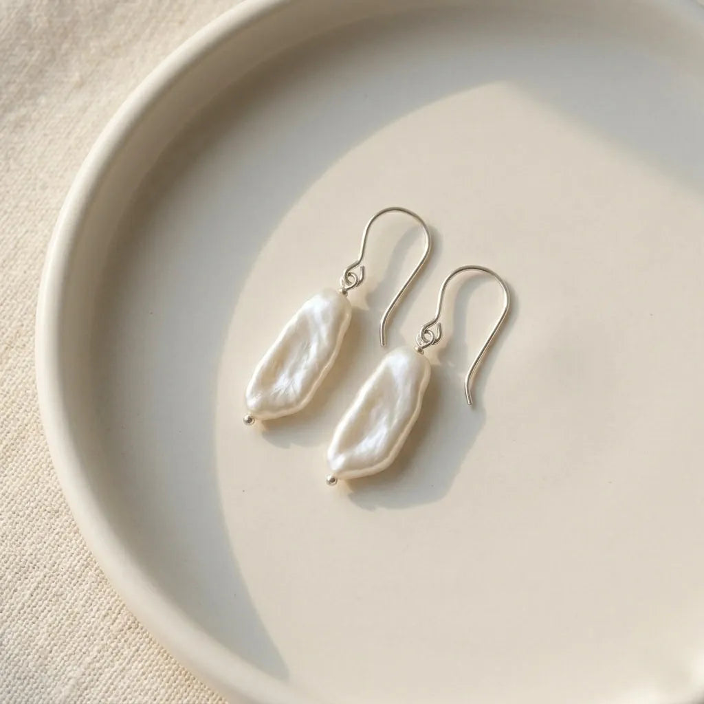 A pair of natural white baroque pearl earrings with silver French hooks, elegantly resting on the edge of a minimalist white ceramic dish.